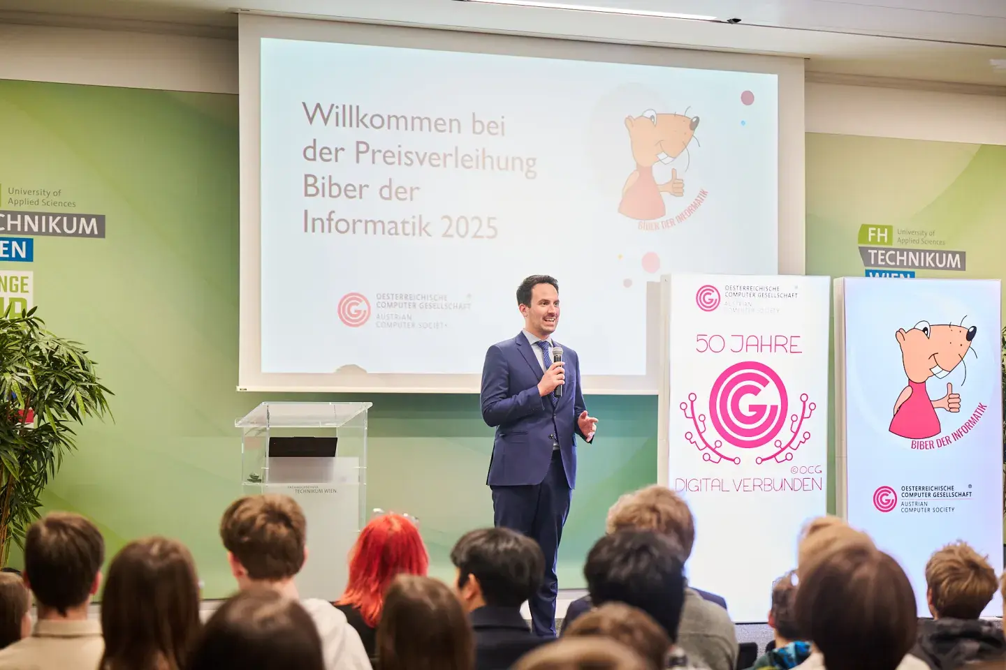 Bildungsminister Christoph Wiederkehr lobte die Gewinner*innen beim Biber der Informatik 2025 (auf der Bühne, Publikum von hinten, volles Haus)