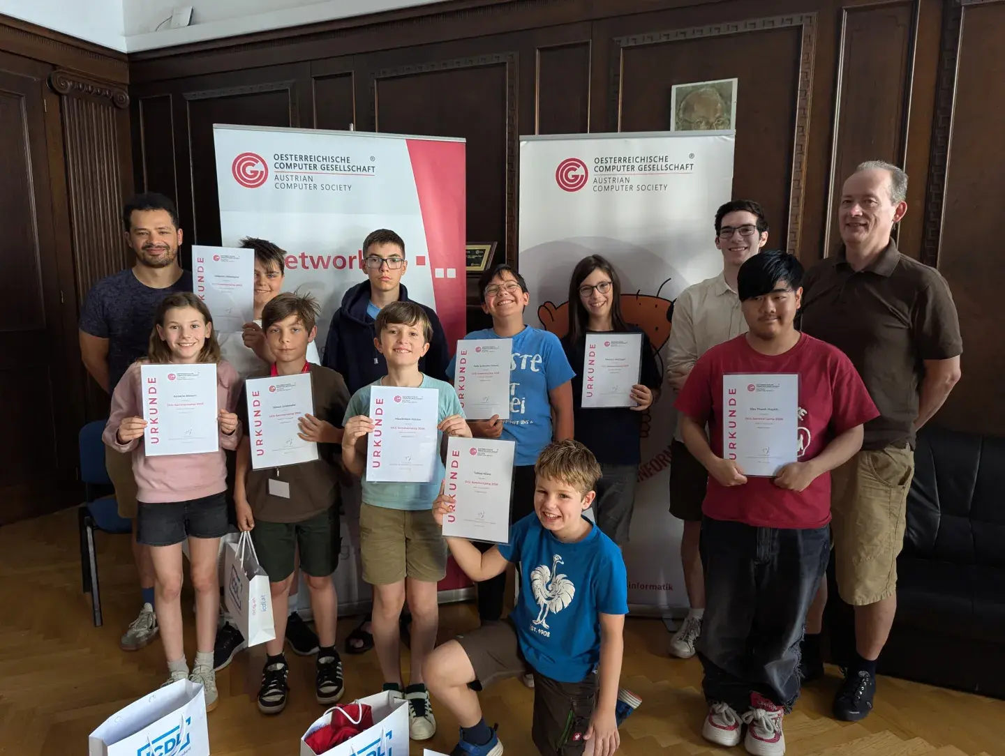 Gruppenfoto: Liam Baumann, 9 TeilnehmerInnen des Sommercamps mit ihren Urkunden, Elias Wiesenhofer, Wilfried Baumann; im Heinz Zemanek Saal, OCG Banner im Hintergrund