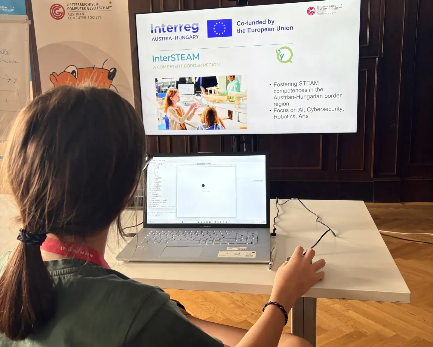 Ansicht von hinten auf ein Mädchen, das programmiert. Vorne Biber der Informatik Roll-up und Interreg Projekt Projeziert auf Bildschirm, OCG Sommercamp, Zemanek Saal