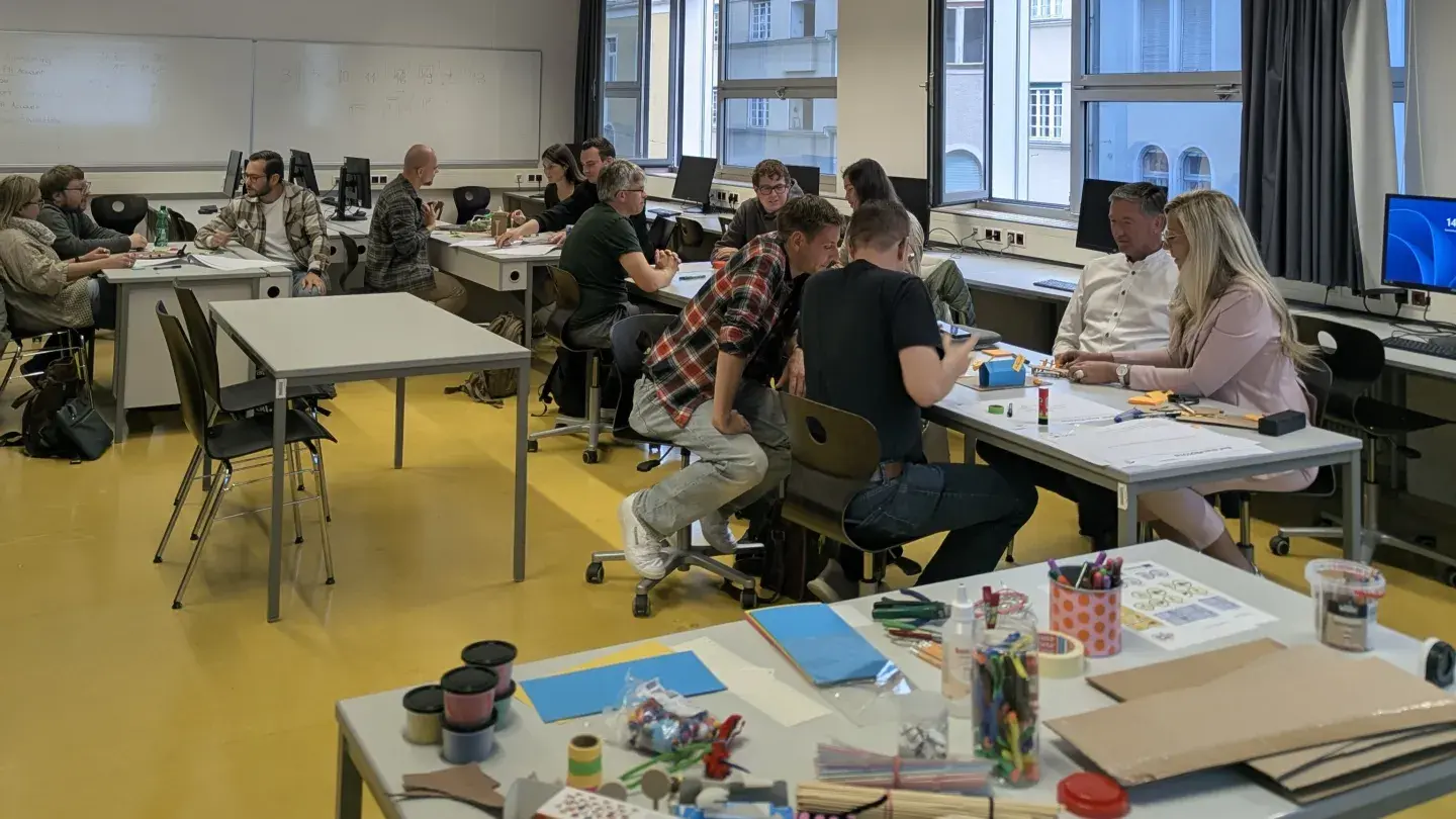 Unterrichtsraum, im Vordergrund ein Tisch mit Lernutensilien, unterschiedliches Material; mehrere Tische mit erwachsenen Personen, die am Workshop Design Thinking (ComeThinkAgain-Projekt) teilnehmen und fokussiert arbeiten, Tag der digitalen Bildung, PH Steiermark (c) OCG