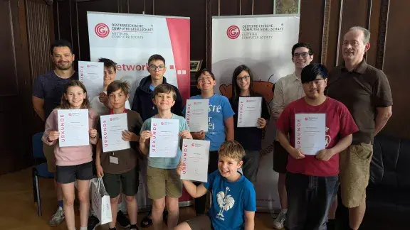 Gruppenfoto: Liam Baumann, 9 TeilnehmerInnen des Sommercamps mit ihren Urkunden, Elias Wiesenhofer, Wilfried Baumann; im Heinz Zemanek Saal, OCG Banner im Hintergrund