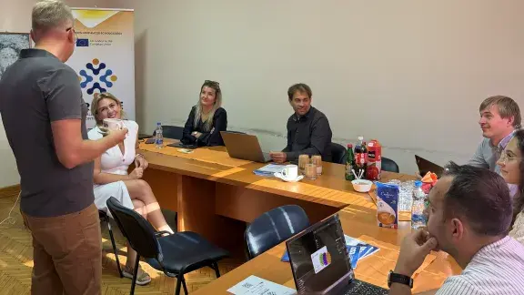 Foto zeigt 7 mit Laptops arbeitende Personen und das ARAISE - Accessible Robotics and AI Supporting Education Logo im Hintergrund beim Projekt-Meeting in Belgrad